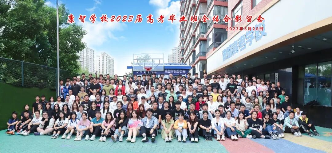康智校园：教学楼、校门招牌与塑胶球场。该照片为 2023 届毕业班合影场景，用于展示校园环境。